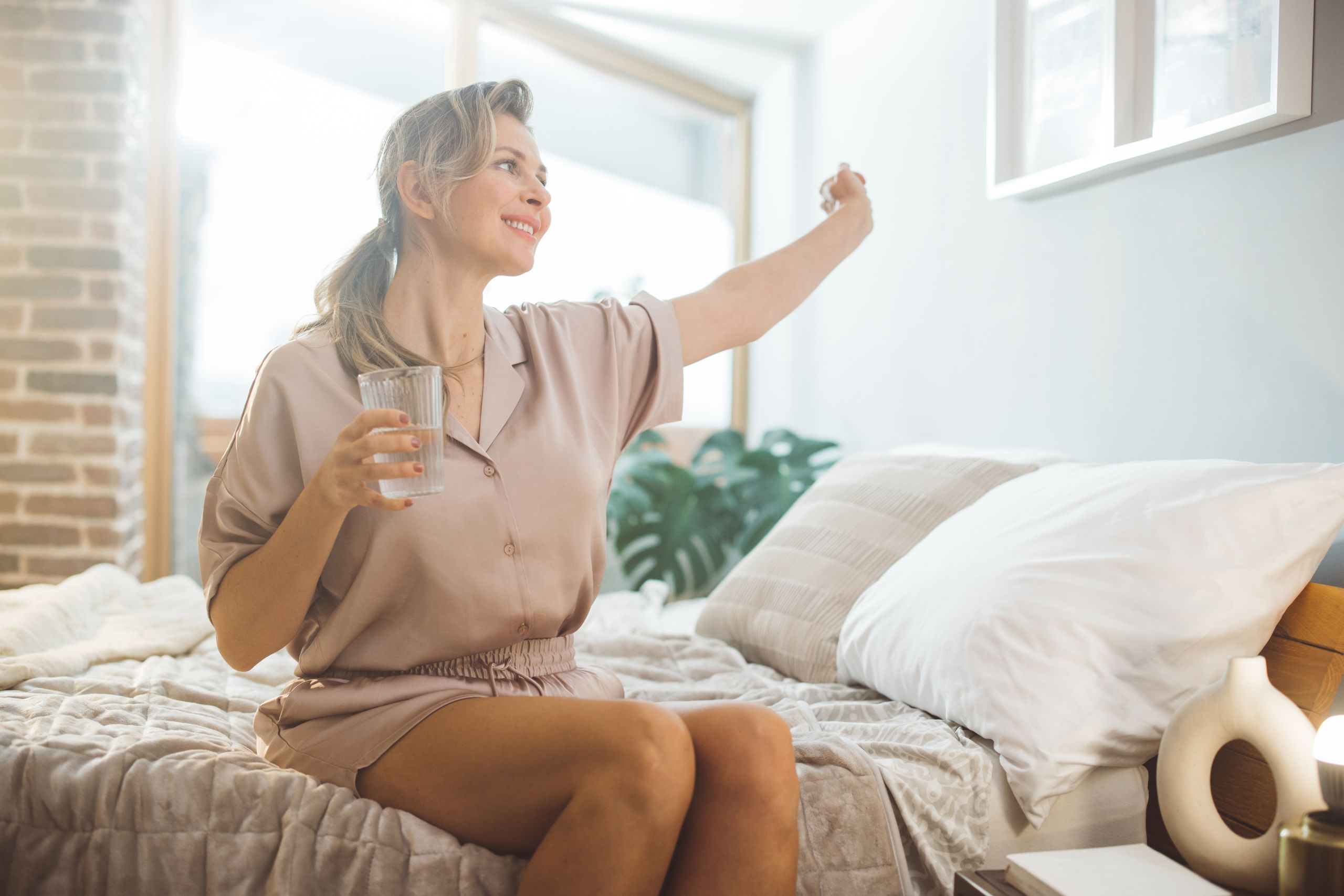 A smiling woman stretching her arms , sitting on her bed as she has just woken up, the sun is shining through the window in her room and she is holding a glass of water